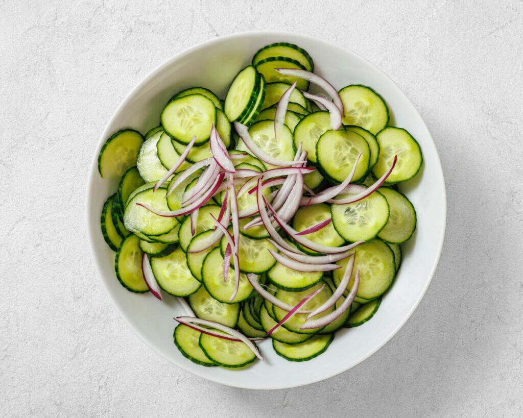 A white bowl filled with sliced cucumbers and thinly sliced red onions on a light, textured surface.
