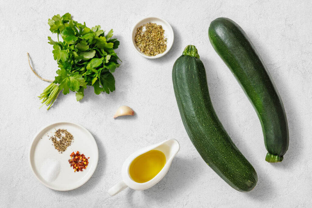 Two zucchinis, fresh parsley, a garlic clove, olive oil, dried oregano, and plates with salt, pepper, and red pepper flakes arranged on a white surface.