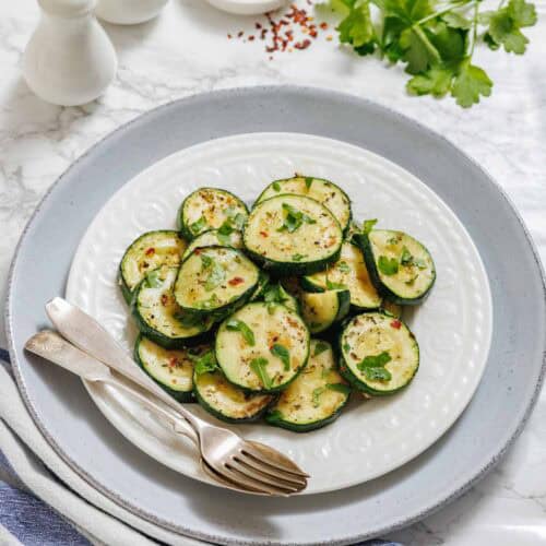 A white plate of sliced, cooked zucchini garnished with herbs, served with a fork and spoon on a gray plate with parsley, red pepper flakes, and salt and pepper shakers nearby.