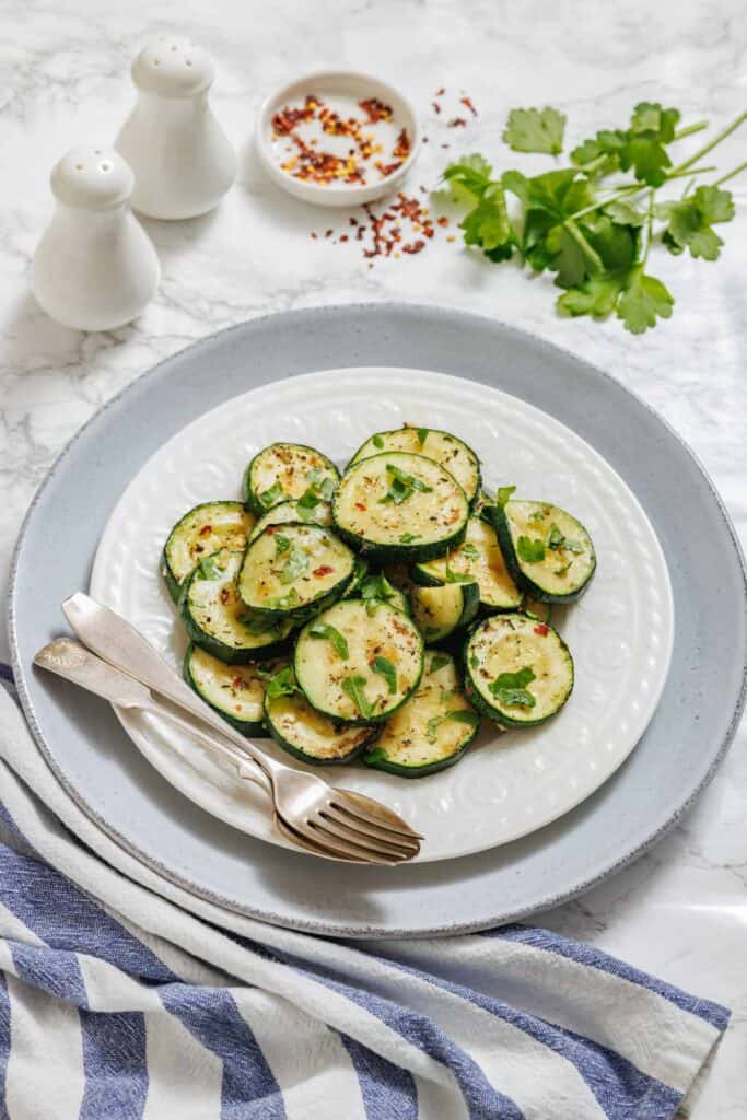 A white plate of sliced, cooked zucchini garnished with herbs, served with a fork and spoon on a gray plate with parsley, red pepper flakes, and salt and pepper shakers nearby.