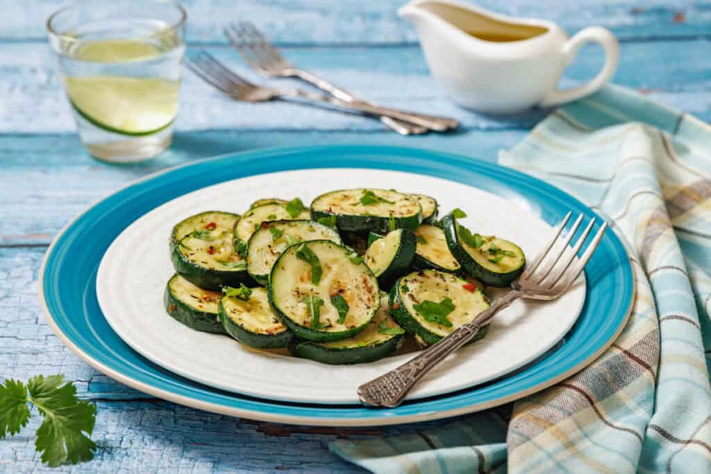 A plate of sliced, grilled zucchini garnished with herbs, placed on a blue and white dish with a fork, napkin, and a small pitcher of dressing in the background.