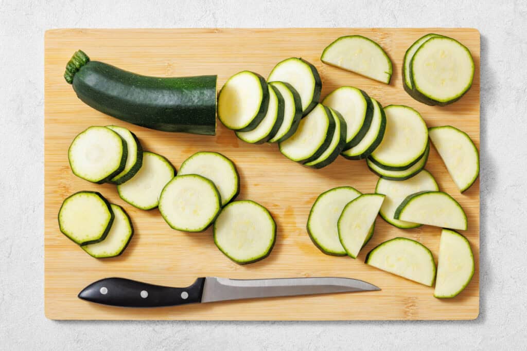A whole zucchini and several slices are arranged on a wooden cutting board with a black-handled knife.