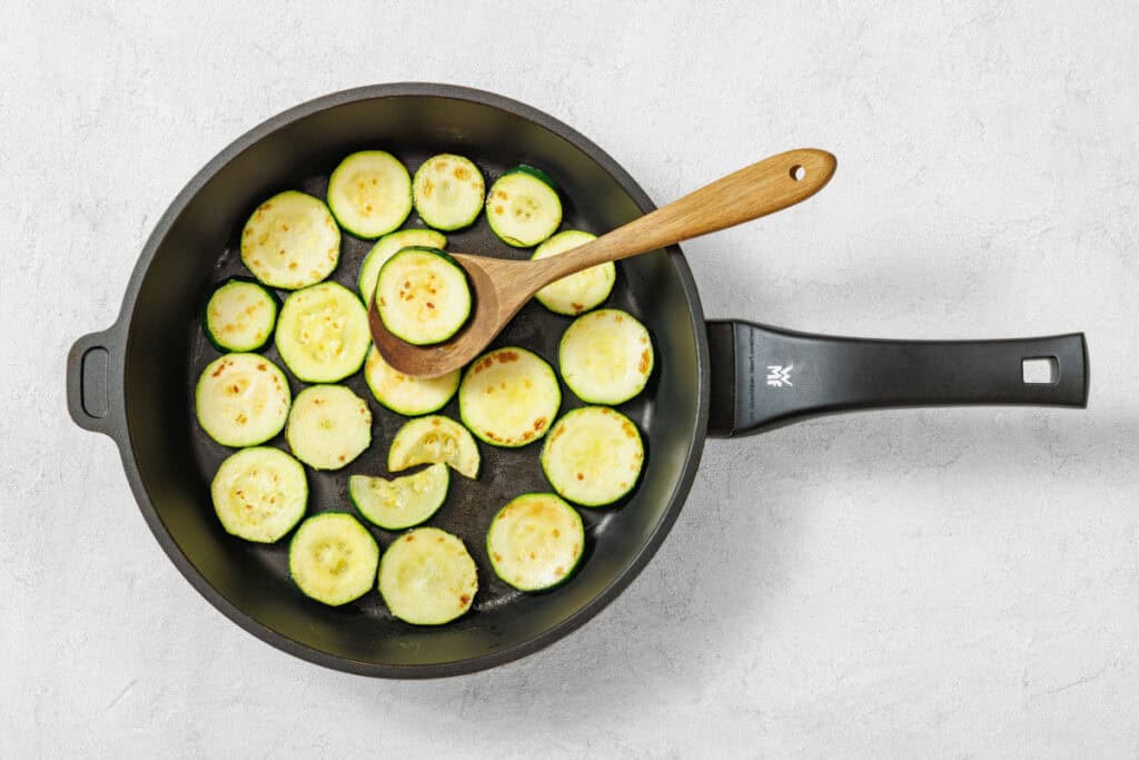 Sliced zucchini rounds being sautéed in a black frying pan with a wooden spoon on a light gray surface.