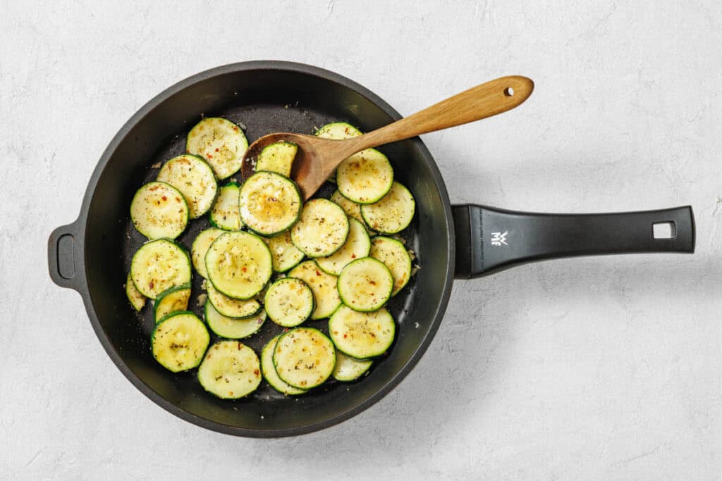 Sliced zucchini being sautéed in a black frying pan with a wooden spoon on a light gray surface.