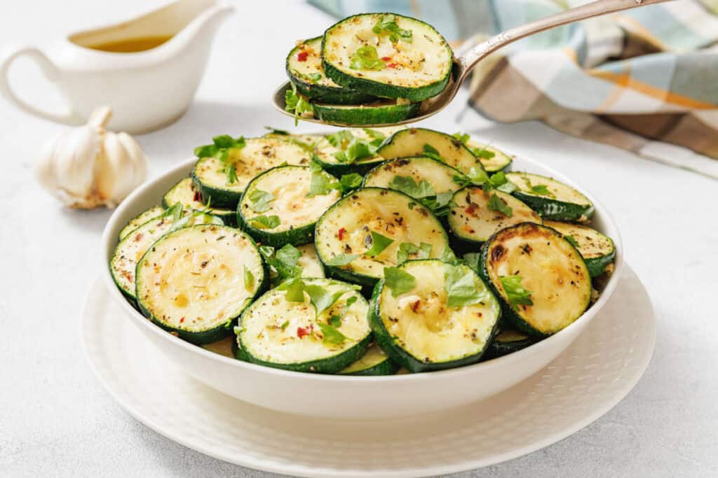 A white bowl filled with sliced, cooked zucchini garnished with herbs, with a spoon lifting some pieces. A sauce boat and garlic bulb are in the background.