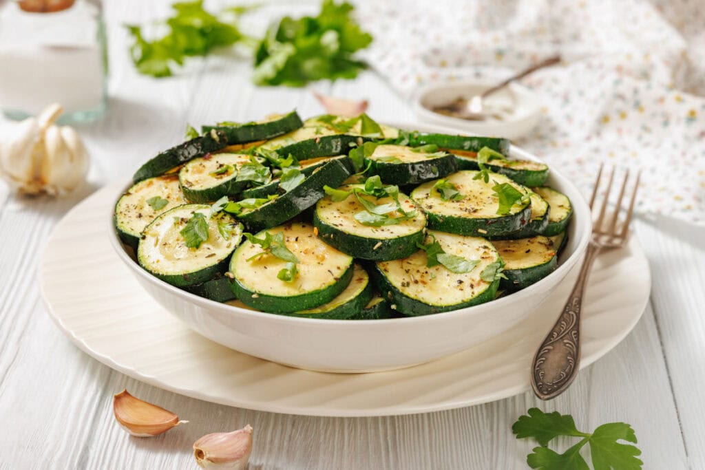 A bowl of sliced, cooked zucchini garnished with chopped parsley, with garlic cloves and herbs scattered on a white wooden table.