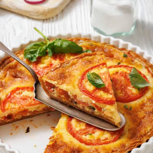 A slice of tomato pie being lifted with a serving utensil from a white fluted dish. A whole tomato pie is visible in the dish, garnished with tomato slices and basil.