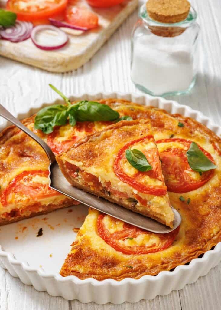 A slice of tomato pie being lifted with a serving utensil from a white fluted dish. A whole tomato pie is visible in the dish, garnished with tomato slices and basil.