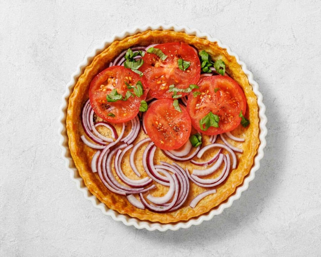 Overhead shot of a tomato tart in a white fluted dish, featuring red onion rings, tomato slices, and herbs.