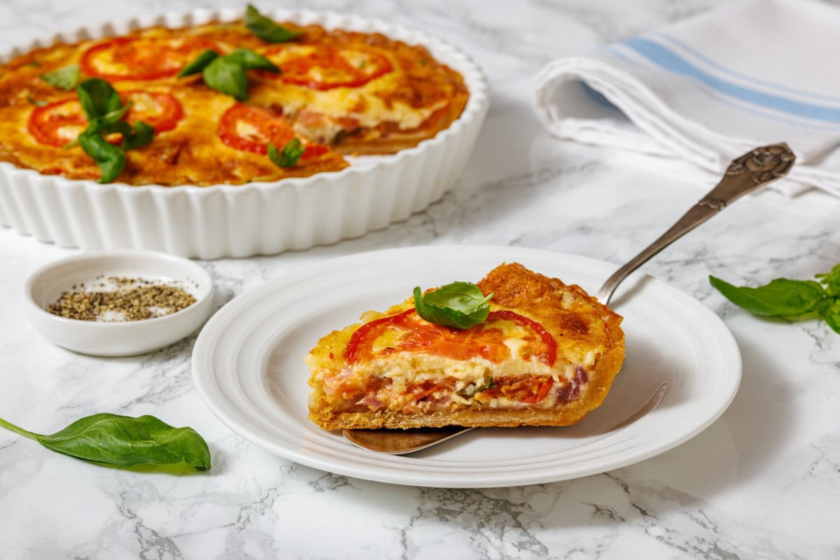 A slice of tomato pie on a white plate with a serving utensil, and a whole tomato pie in a white fluted dish, are shown on a marble surface with a small bowl of spices and basil leaves.