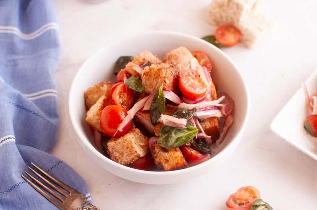 Overhead shot of panzanella salad in a white bowl, featuring toasted bread cubes, cherry tomatoes, red onion, and basil leaves, with a fork and cloth.