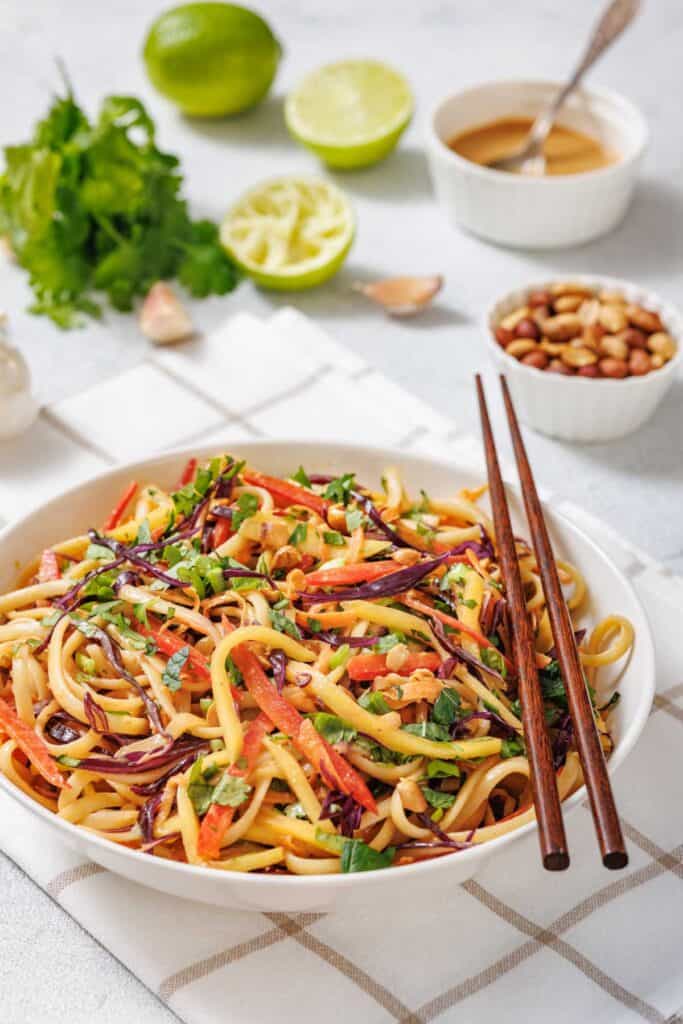 A bowl of colorful noodle salad with vegetables, garnished with herbs and chopped peanuts, chopsticks resting on the bowl. Lime, cilantro, peanuts, and sauce are in the background.