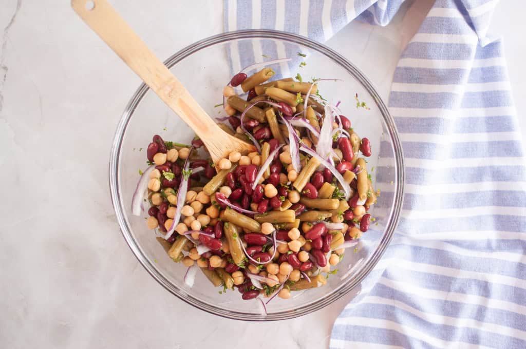 A glass bowl filled with a mixed bean salad including kidney beans, chickpeas, green beans, and sliced red onion, with a wooden spoon and a striped cloth nearby.