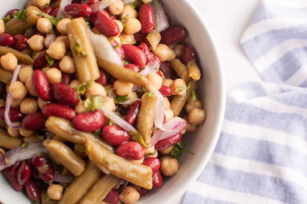A bowl of three bean salad with kidney beans, chickpeas, green beans, and sliced red onions next to a blue and white striped cloth.