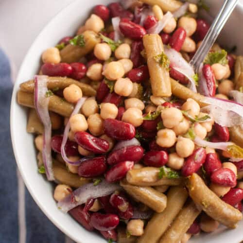 A bowl of three bean salad with chickpeas, kidney beans, green beans, sliced red onions, and chopped herbs, with a fork inside the bowl.