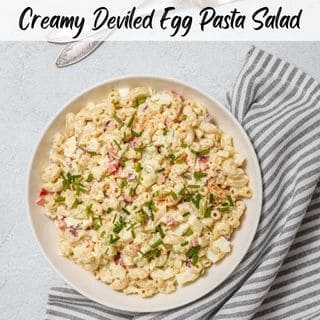 A creamy deviled egg pasta salad served in a white bowl, garnished with chopped chives and paprika, placed on a striped napkin with vintage forks beside it.