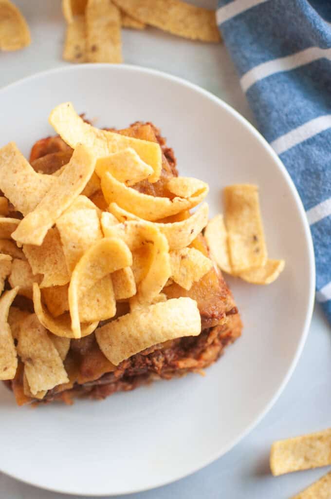 A close up shot of white plate with a serving of Frito pie, topped with corn chips, is shown on a light surface.