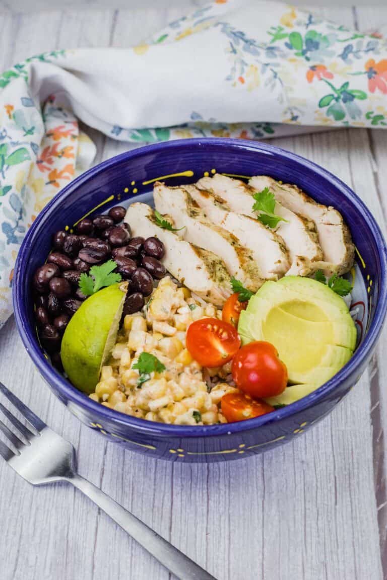 A blue bowl filled with a street corn chicken rice bowl, featuring grilled chicken, corn, tomatoes, avocado, and black beans, is shown with a fork and cloth.