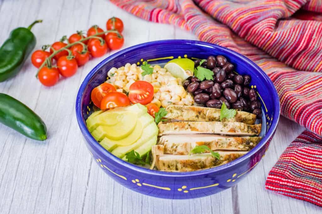 A blue bowl filled with a street corn chicken rice bowl, featuring grilled chicken, corn, tomatoes, avocado, and black beans, is shown with vegetables and a colorful cloth.