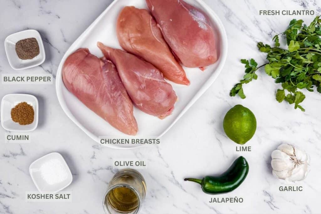 Raw chicken breasts on a white plate with fresh cilantro, lime, jalapeño, garlic, olive oil, cumin, black pepper, and kosher salt arranged on a white marble surface.