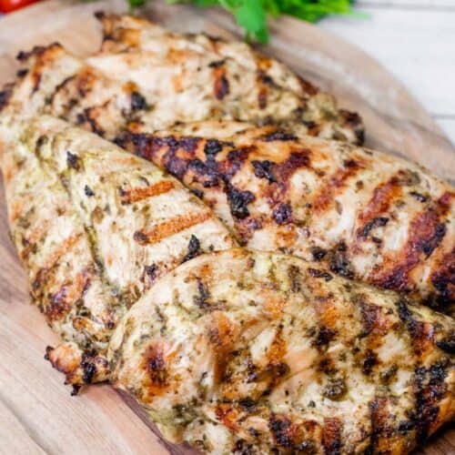 Close-up of grilled chicken on a wooden board with cilantro and tomatoes in the background.