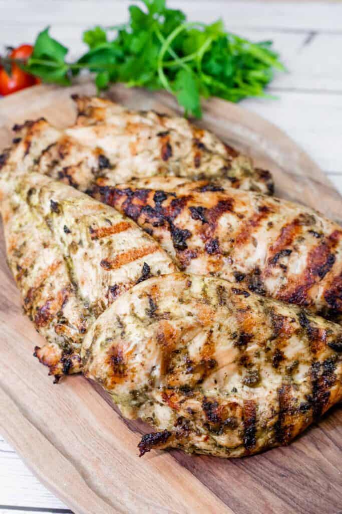 Close-up of grilled chicken on a wooden board with cilantro and tomatoes in the background.