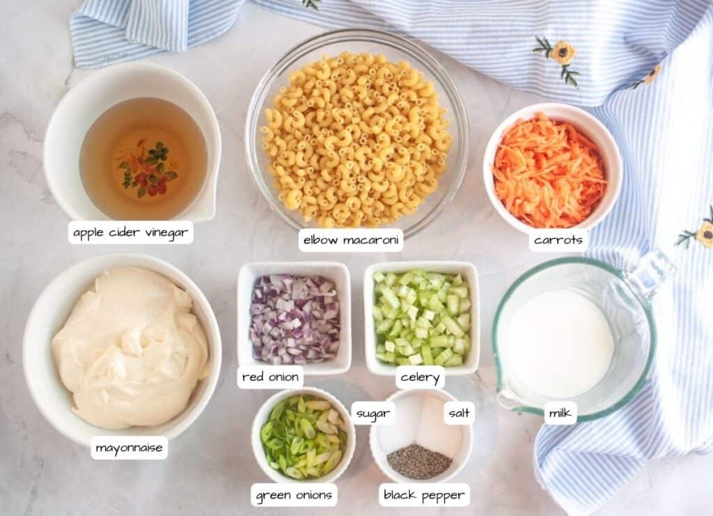 Overhead shot of ingredients for Hawaiian macaroni salad, including macaroni, carrots, mayonnaise, onions, celery, milk, seasonings, and vinegar, arranged on a light surface with a striped cloth and labels for each ingredient.