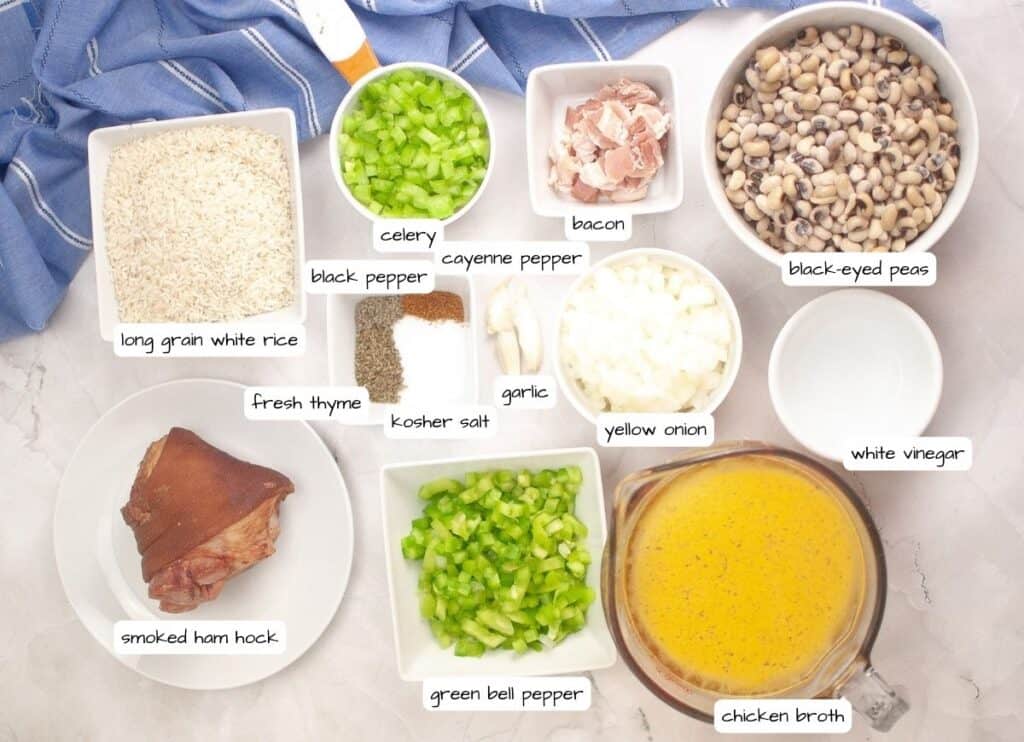 Ingredients for Hoppin’ John are arranged on a countertop, including rice, black-eyed peas, bacon, vegetables, spices, a smoked ham hock, chicken broth, and vinegar.