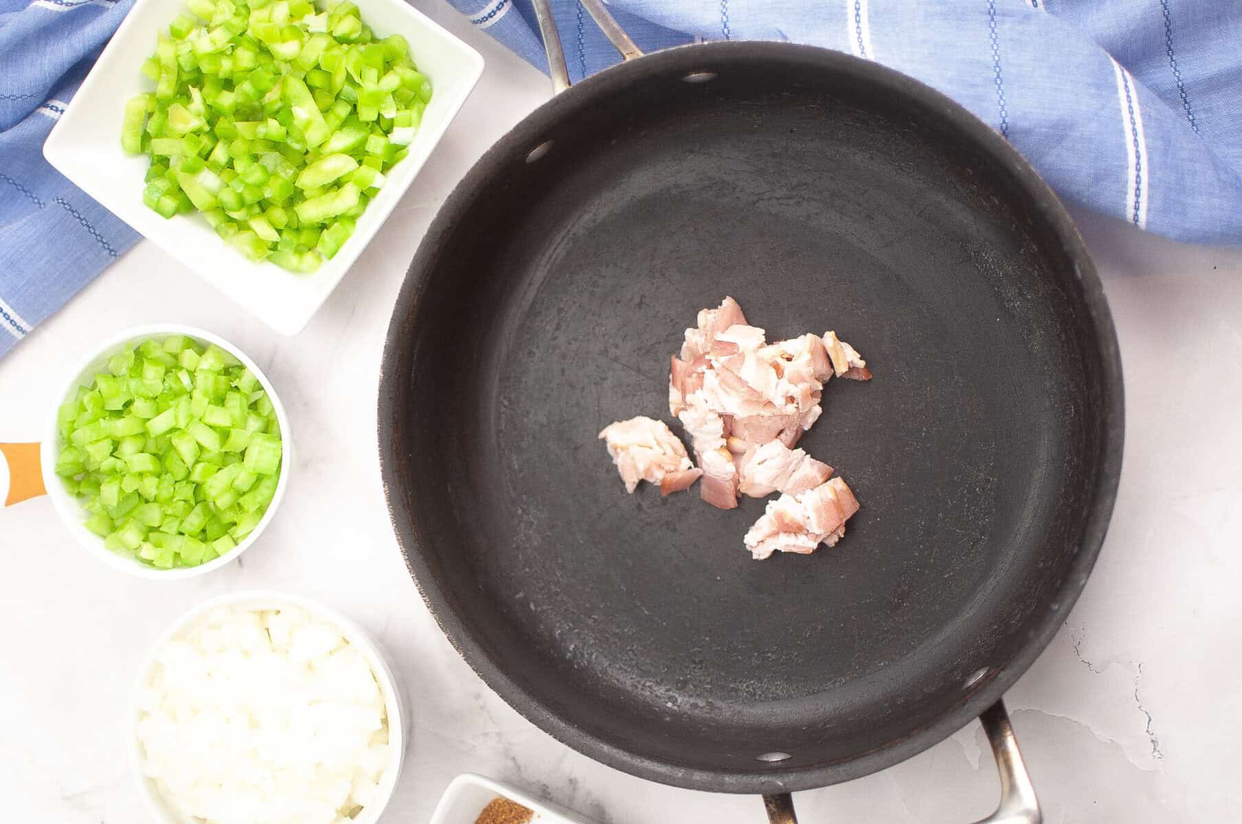 A skillet with chopped raw bacon in the center, surrounded by bowls of diced green bell pepper, celery, and onion on a countertop with a blue cloth.