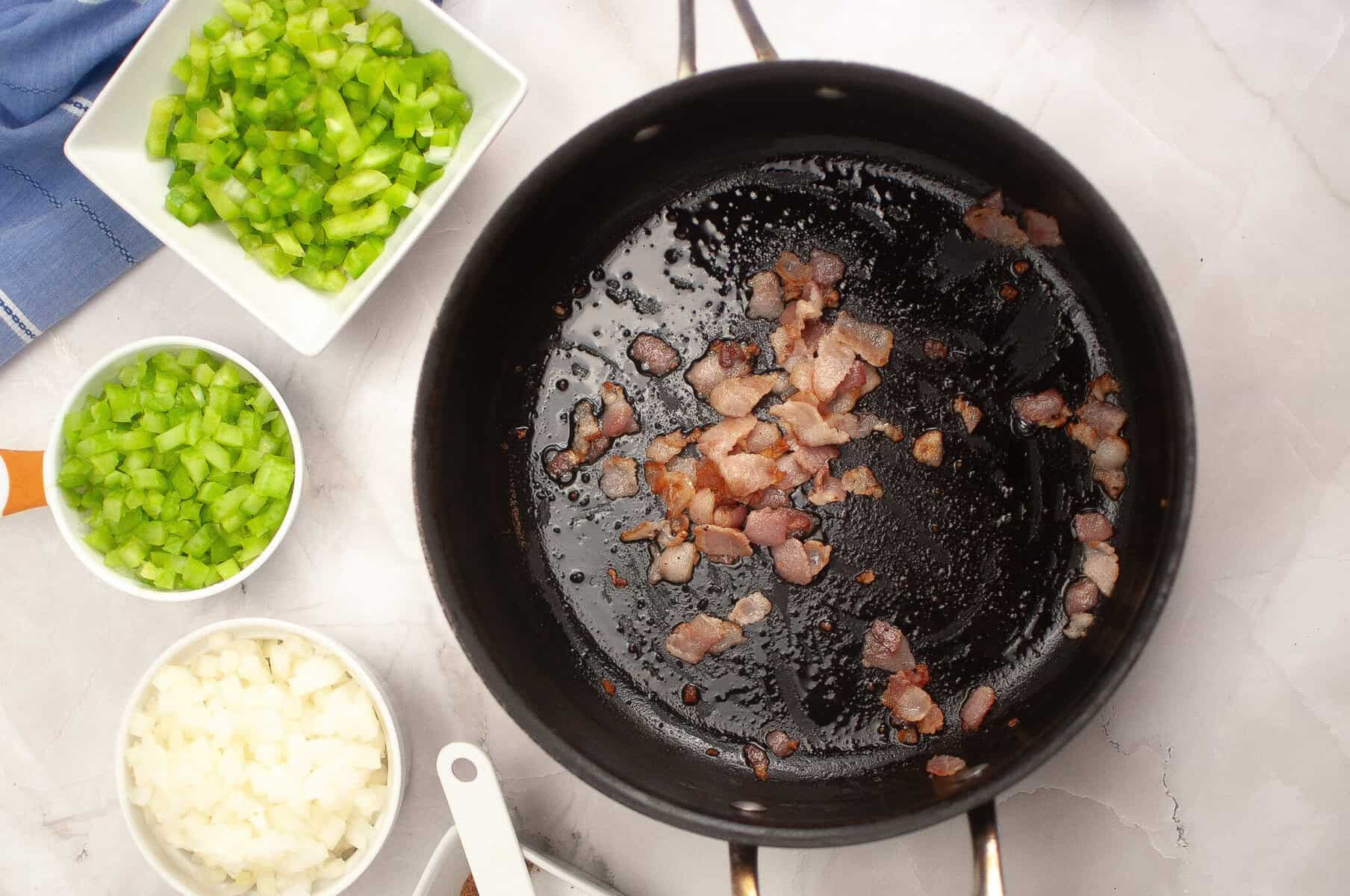 Chopped bacon cooking in a black pan with diced onions, green bell peppers, and celery in bowls nearby on a marble countertop.