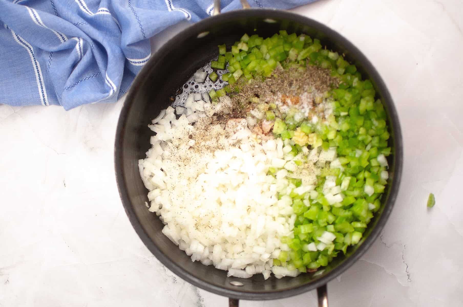Chopped onions and green bell peppers with seasoning in a black pot, ready to be cooked, on a white countertop with a blue towel nearby.