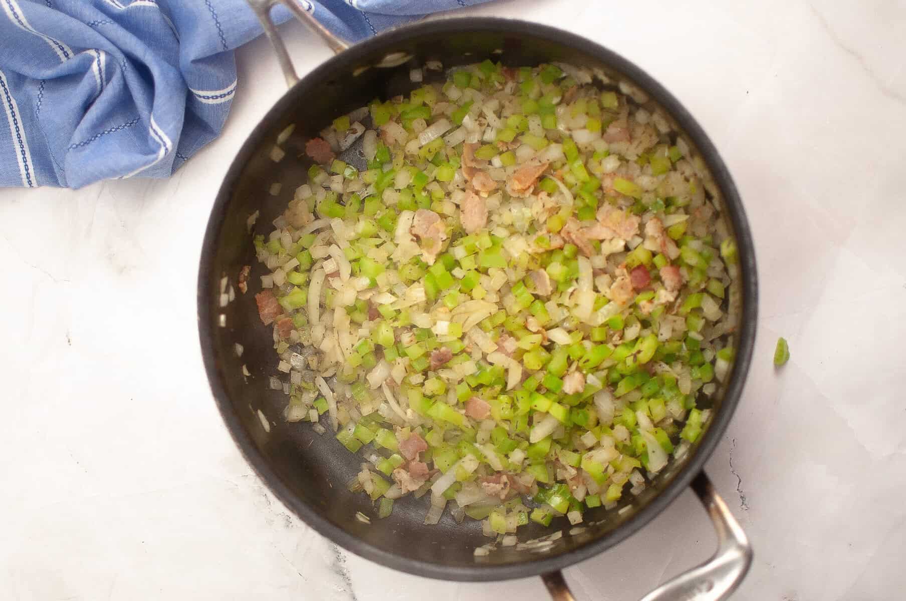 Chopped celery, onions, and bacon pieces are sautéing in a black pot on a white marble surface, with a blue kitchen towel nearby.