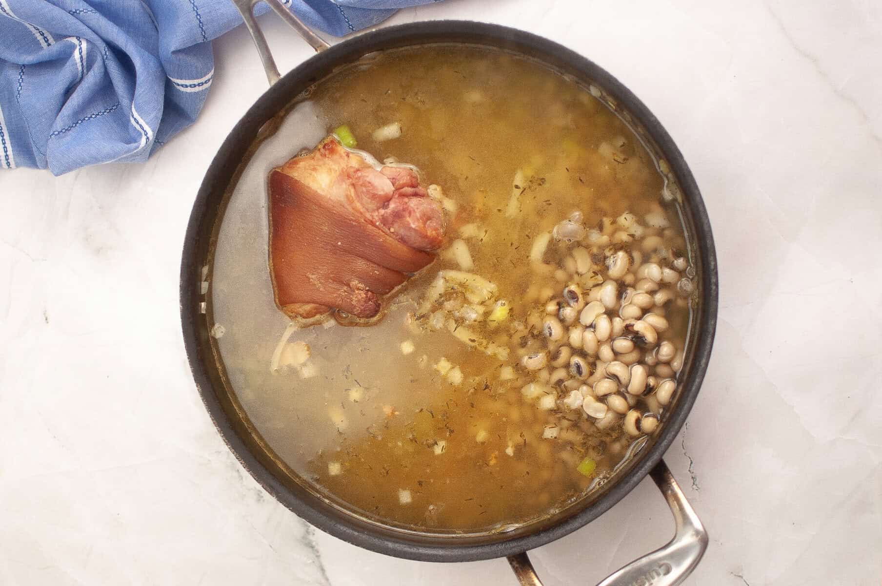 A pot of soup with a ham hock and black-eyed peas simmering in broth, placed on a white marble surface next to a blue cloth.