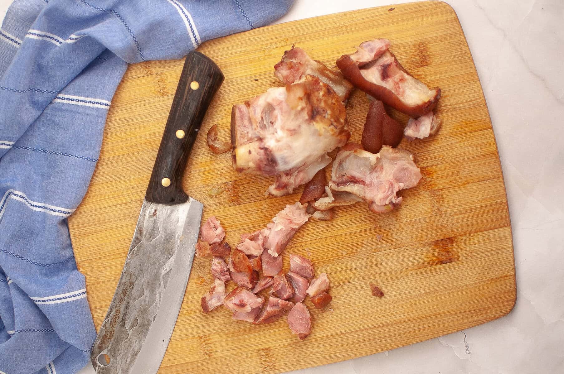 Chopped smoked ham hock on a wooden cutting board with a large knife and blue kitchen towel.