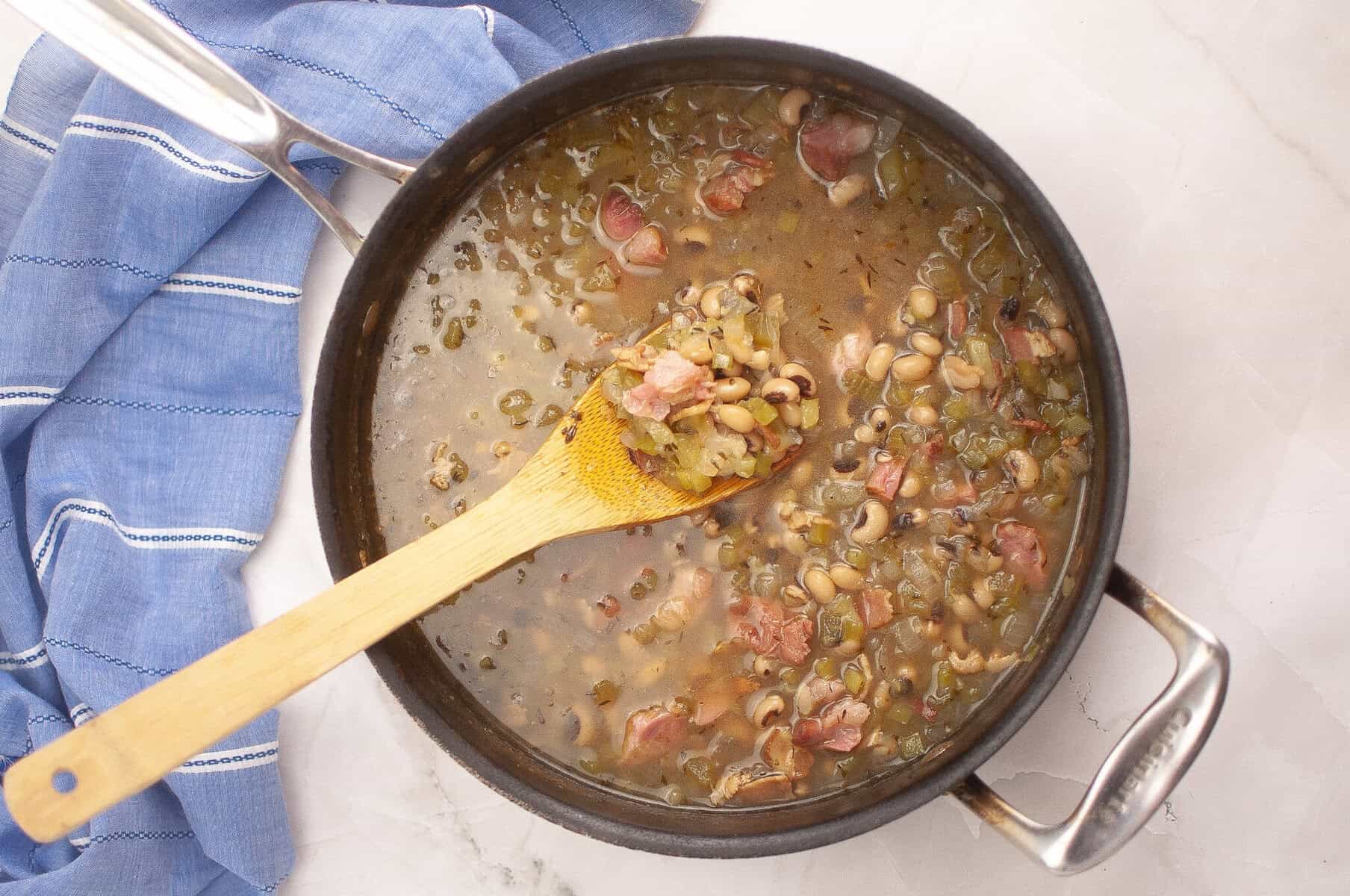 A pot of cooked black-eyed peas with ham and vegetables, stirred with a wooden spoon, sits on a marble countertop next to a blue striped cloth.