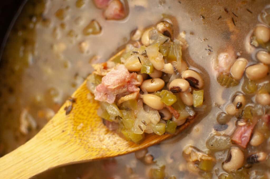 A wooden spoon holds a serving of black-eyed peas, diced vegetables, and pieces of ham in a broth.