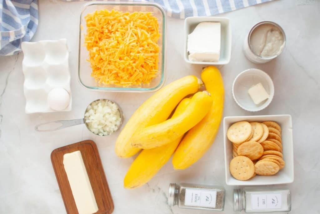 Ingredients for a casserole arranged on a countertop, including yellow squash, shredded cheese, cream cheese, butter, egg, onion, crackers, seasonings, and a small can of soup.