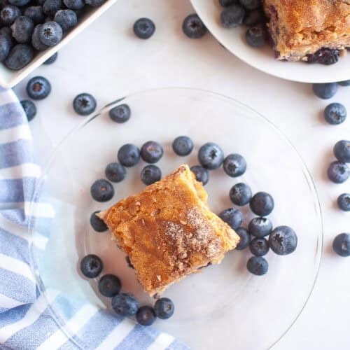 A square piece of Blueberry Boy Bait on a clear plate surrounded by blueberries, with a bowl of blueberries and more cake on a separate plate nearby.