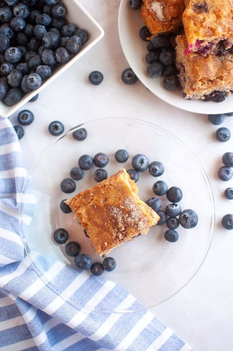 A square piece of Blueberry Boy Bait on a clear plate surrounded by blueberries, with a bowl of blueberries and more cake on a separate plate nearby.