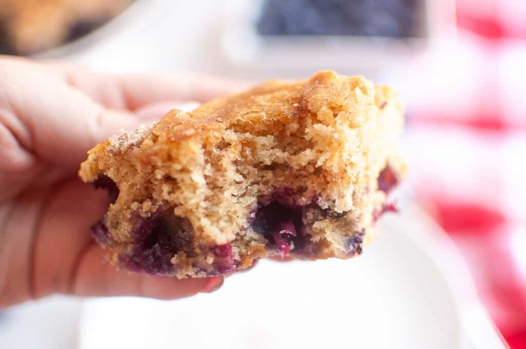 A hand holds a partially eaten piece of blueberry coffee cake, showing a soft, crumbly texture and visible blueberries inside.