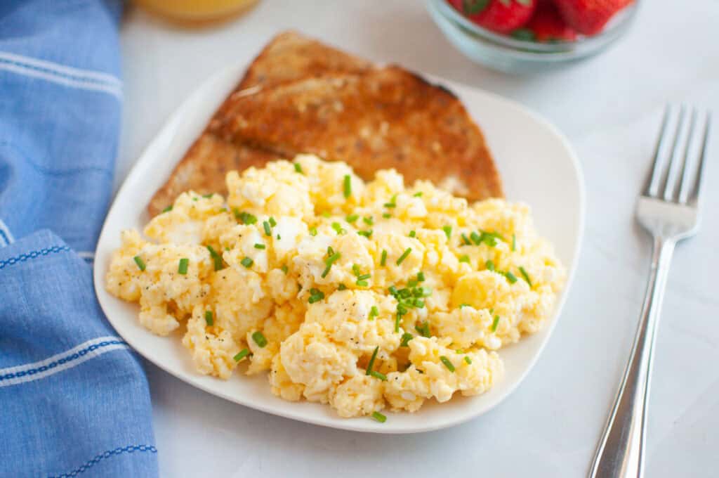 A plate of scrambled eggs garnished with chives, served with two slices of toast. A fork, blue napkin, and bowl of strawberries are nearby.