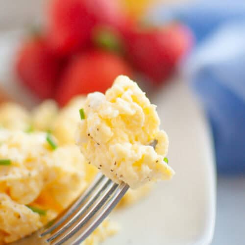 A fork holds a bite of scrambled eggs, with strawberries and a glass of orange juice blurred in the background on a breakfast table.