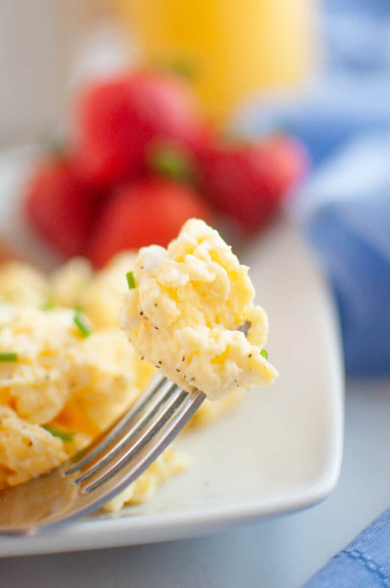 A fork holds a bite of scrambled eggs, with strawberries and a glass of orange juice blurred in the background on a breakfast table.