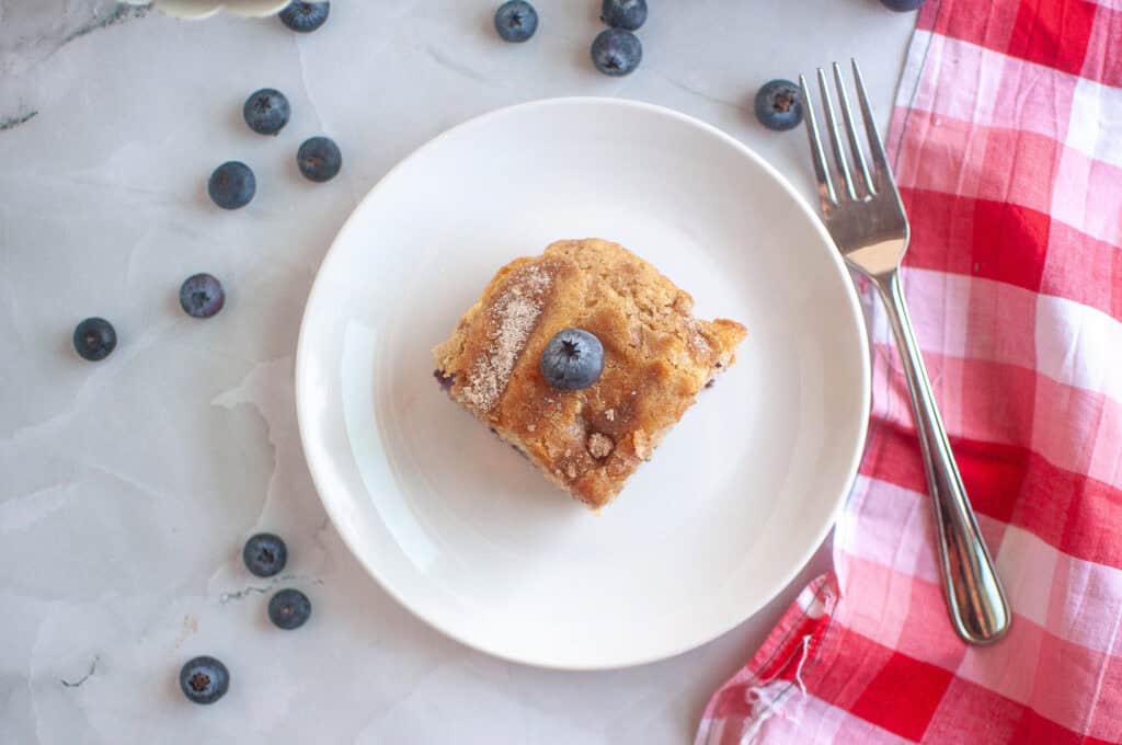 A slice of blueberry coffee cake on a white plate with a fork, garnished with a blueberry, surrounded by loose blueberries and a red checkered napkin.
