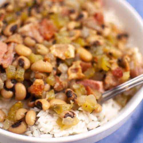 A bowl of white rice topped with black-eyed peas, chopped vegetables, and pieces of bacon, with a spoon resting inside.