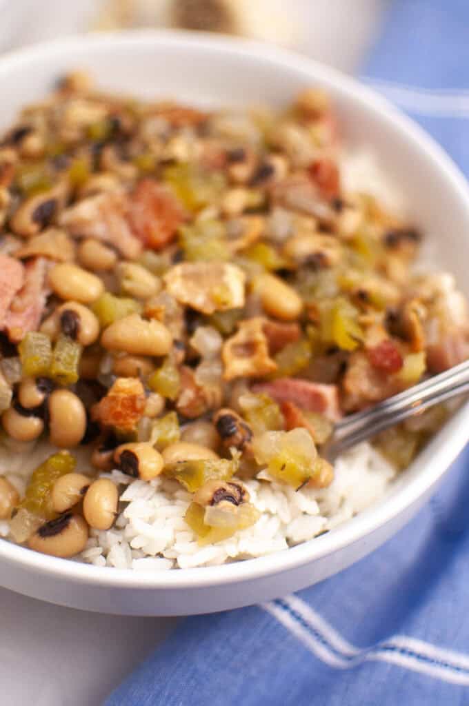 A bowl of white rice topped with black-eyed peas, chopped vegetables, and pieces of bacon, with a spoon resting inside.