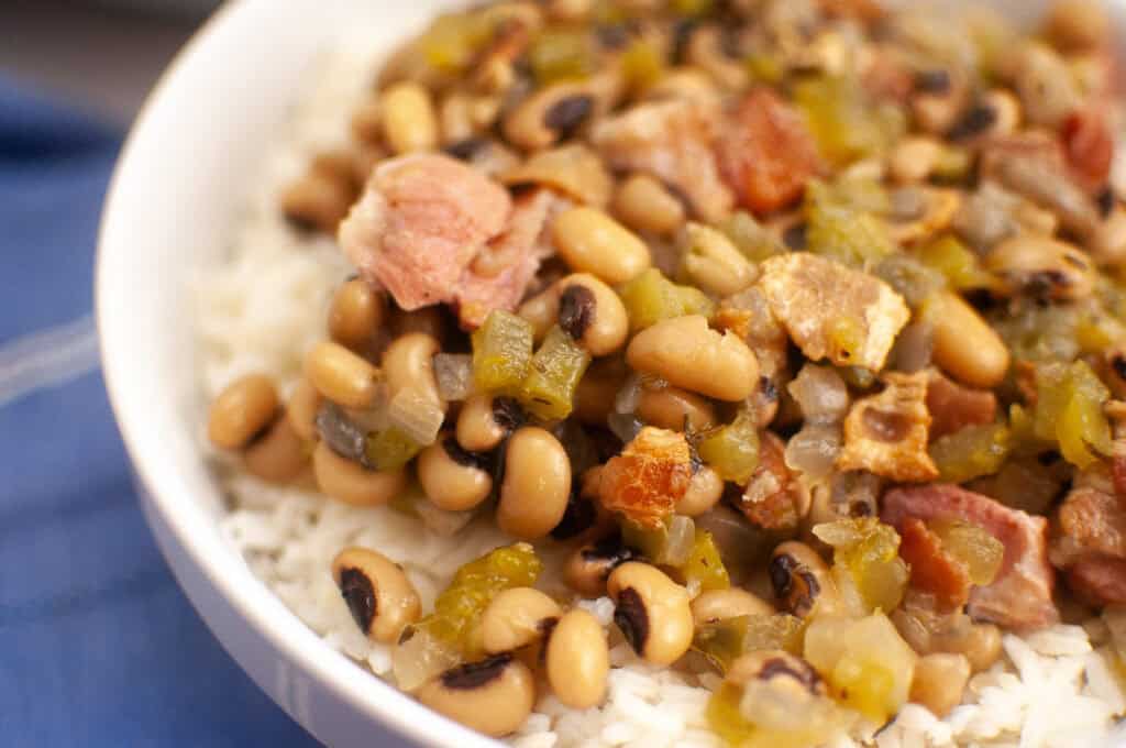 A close-up of a bowl filled with white rice topped with black-eyed peas, chopped green peppers, onions, and pieces of bacon.