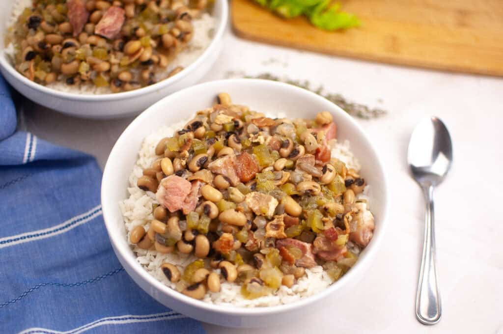 A bowl of white rice topped with a mixture of black-eyed peas, chopped ham, bacon, and vegetables sits next to a spoon on a white surface.