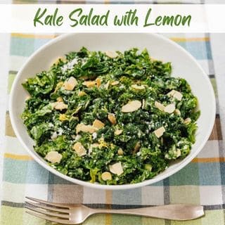 A white bowl of kale salad topped with sliced almonds and grated cheese sits on a checkered tablecloth with a fork beside it. Text at the top reads "Kale Salad with Lemon.
