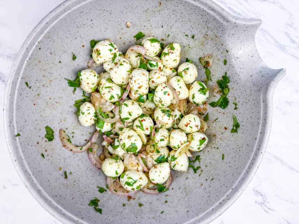 A bowl of marinated mozzarella balls mixed with sliced red onions and chopped herbs, seasoned with spices, on a white marble surface.
